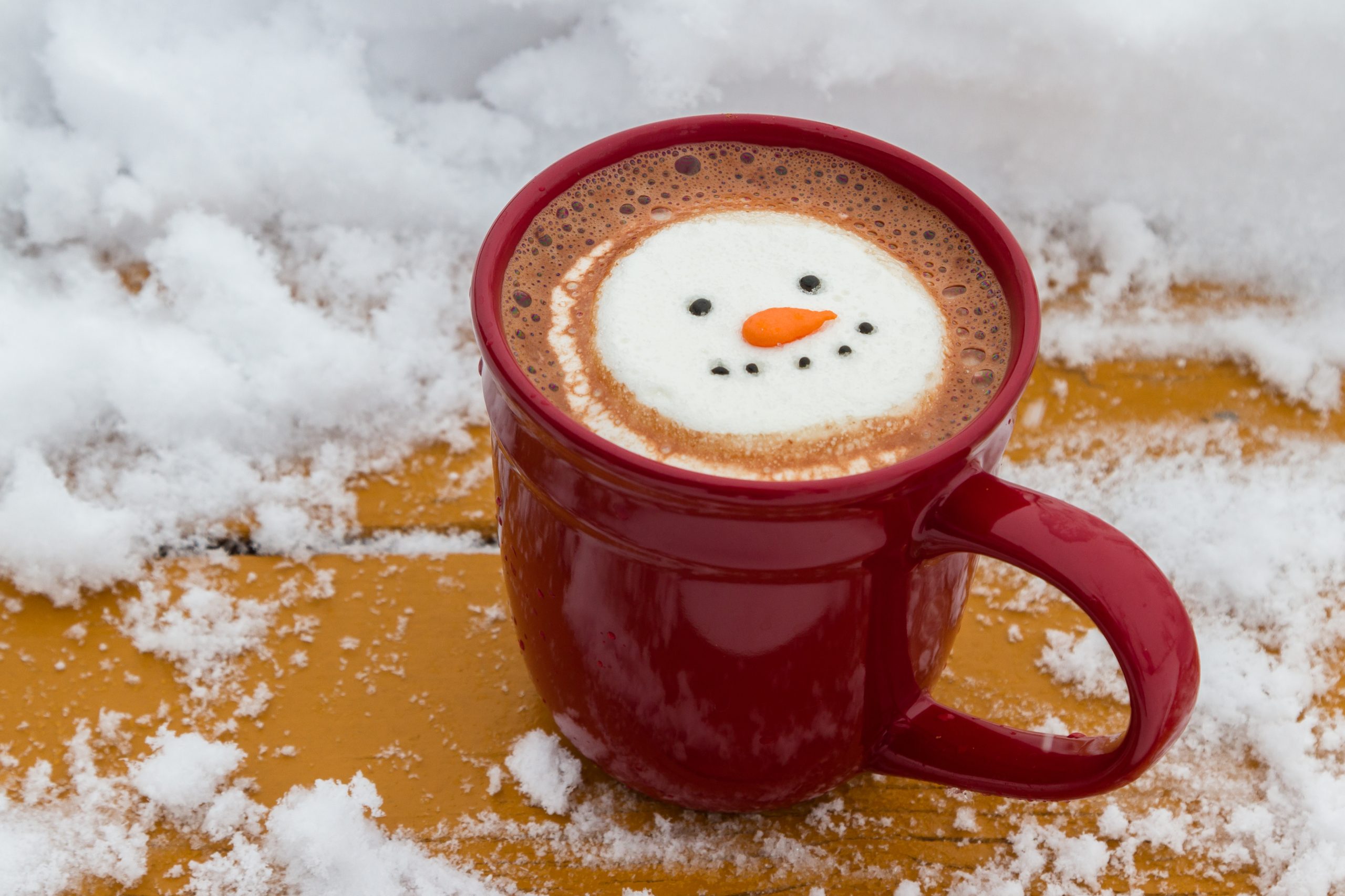 A red mug of hot cocoa on a snowy table with a smiling snow-person marshmallow