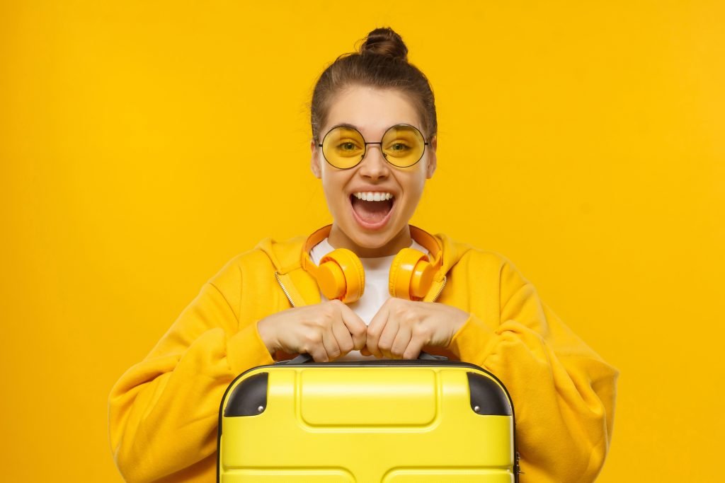 Happy excited teenage girl wearing hoodie, colored glasses and headphones around neck, holding travel suitcase, feeling ready for flight, isolated on yellow background