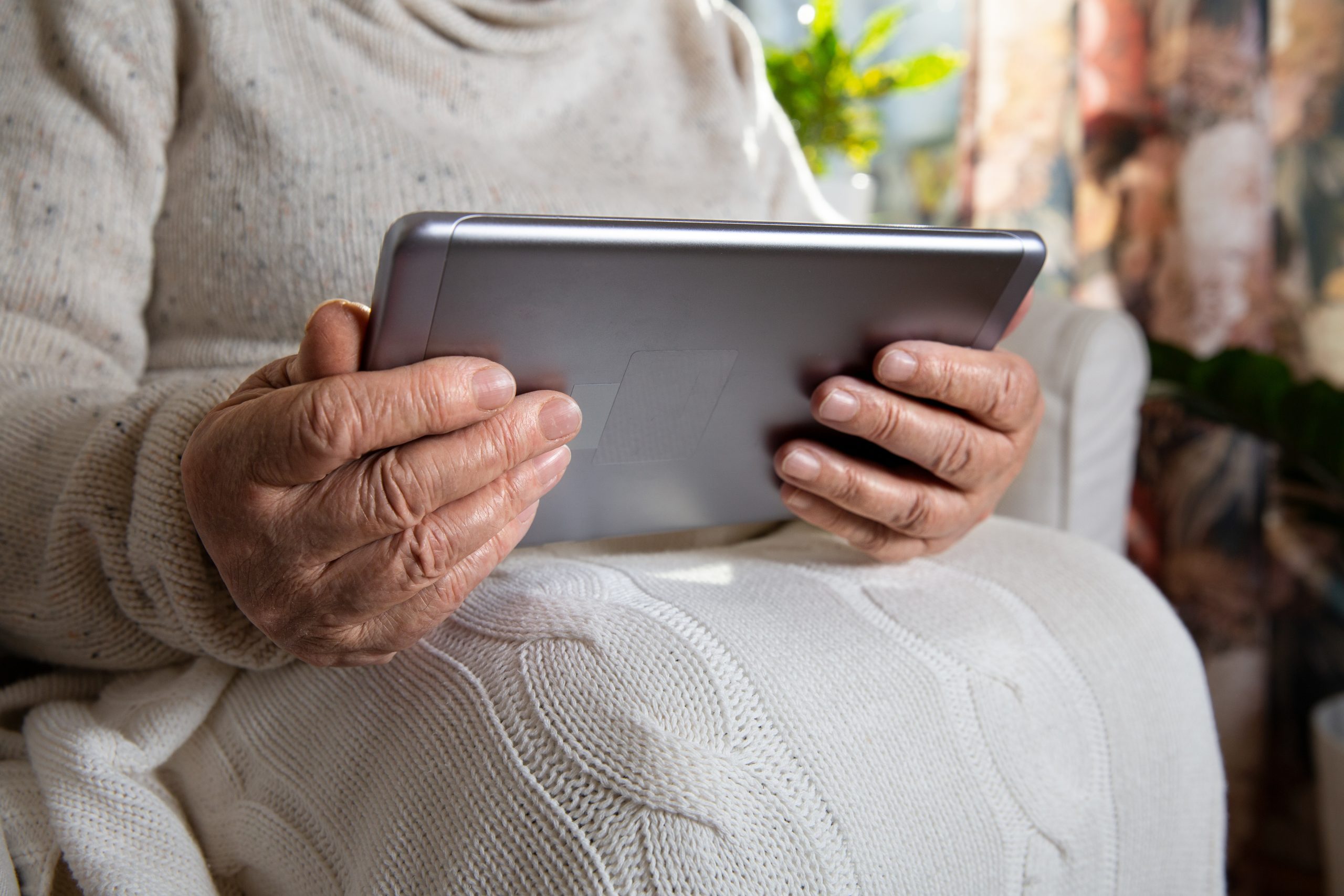 Close-up wrinkled hands of old woman with tablet computer. Senior lady sitting in chair with blanket. Warm and cozy, time to relax, browsing, with tablet in hand
