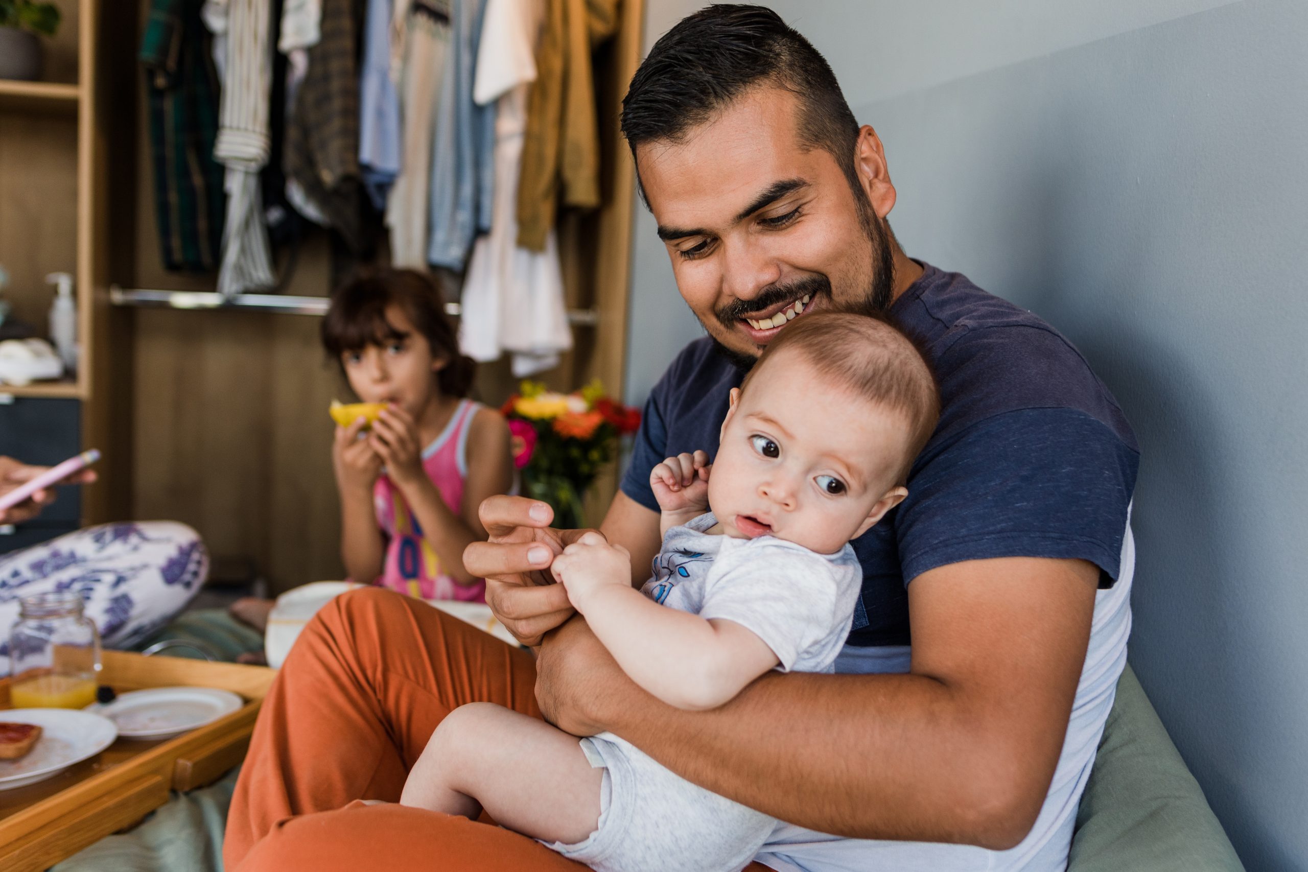 A father lovingly holding his infant while smiling. The father and infant are in a room with another child in the background.